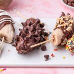 Four frozen chocolate covered banana halves on sticks are adorned with an assortment of toppings: nuts, chocolate chips, and vibrant sprinkles. They're artfully arranged on a white platter against a lively pink backdrop.