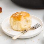 A freshly baked roll with sesame seeds on a white plate and fork, surrounded by honey and more rolls.