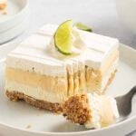 A slice of key lime lush with a graham crust, creamy filling, and zesty lime topping rests on a plate. A fork with a piece of dessert lies invitingly beside it, while a bowl overflowing with fresh limes sits in the background.