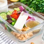 This glass container is perfect for meal prep, featuring a meal prep Greek salad with feta, olives, tomatoes, cucumbers, red onions, and chickpeas. A dressing cup accompanies this convenient dish. Parsley in the background.
