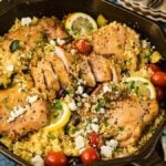 A skillet filled with Mediterranean chicken thighs on a bed of couscous, garnished with cherry tomatoes, lemon slices, crumbled cheese, and herbs.