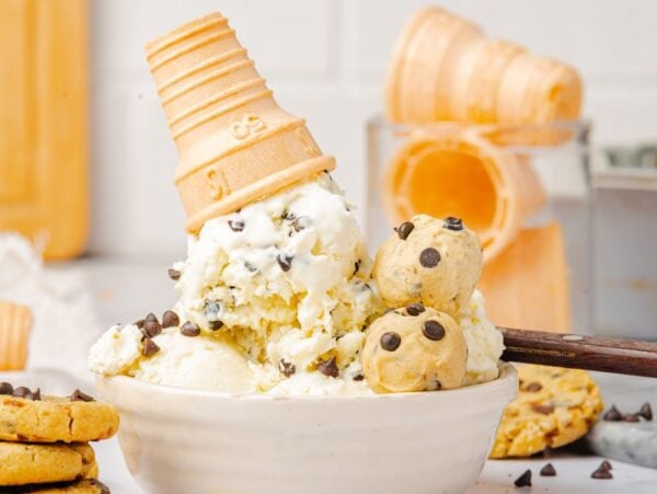 Vanilla no churn cookie dough ice cream in a bowl with a tipped cone, rich chocolate chip cookie dough, and chocolate chip cookies.
