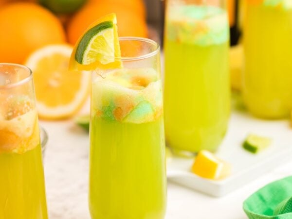 Four glasses of citrus punch with lime and orange garnishes; oranges and a napkin in the background.