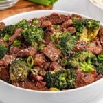 A bowl of slow cooker beef and broccoli stir-fry garnished with sesame seeds and chopped green onions, placed on a table.