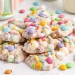 A pile of white chocolate candies with colorful sprinkles and candy pieces, surrounded by various Easter candies and a mug in the background, evokes the cozy charm of a slow cooker treat waiting to be savored.