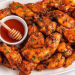 A plate of slow cooker hot honey wings garnished with herbs, served with a small dish of honey and a honey dipper.