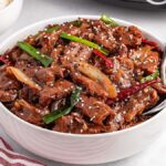 A bowl of stir-fried slow cooker Mongolian beef with green onions and sesame seeds rests on a white surface, accompanied by rice and chopped green onions.