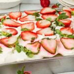 A rectangular strawberry tres leches cake sits in a metal baking pan, topped with white frosting and sliced strawberries.