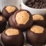 Chocolate-covered peanut butter balls (a classic Buckeyes recipe) are artfully arranged on a surface with a small bowl of chocolate chips in the background.