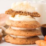 Stack of carrot cake cookies with luscious cream cheese frosting, artfully topped with half a cookie. In the background, fresh carrots, a hint of cinnamon, and a splash of milk complete the scene.