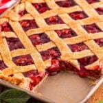 A cherry slab pie with a lattice crust rests in a baking dish, showcasing the missing slice.
