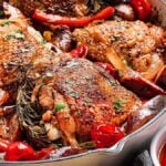 Close-up of a skillet with sumptuous Chicken Scarpariello, featuring roasted chicken, red bell peppers, cherry tomatoes, and a rosemary garnish. A small bowl of cherry tomatoes sits enticingly beside it.