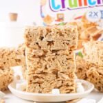A stack of cinnamon marshmallow treats sits elegantly on a white plate, with scattered marshmallows and a Cinnamon Toast Crunch box subtly peeking from the background, adding a nostalgic touch.
