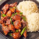 A black plate presents two mounds of white rice alongside a serving of Five Spice Chicken Stir Fry, featuring glazed chicken pieces garnished with sesame seeds and green onions. Nearby, chopsticks await their delicious task.