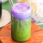 A dreamy iced lavender matcha latte, with vibrant green matcha at the base and a delicate purple froth on top, sits elegantly in a glass with a straw on a wooden coaster. A teapot adds charm to the background.