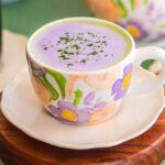 A floral cup cradles a Lavender Matcha Latte, its purple hue complemented by green sprinkles, all resting on a pristine white saucer and wooden coaster.