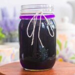 A glass jar filled with dark purple liquid, reminiscent of a lavender simple syrup recipe, is topped with a string-tied lid and placed on a wooden surface. Blurred plants and crockery create an inviting background.