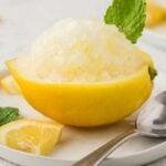 Lemon granita served in a hollowed lemon half, garnished with a mint leaf and lemon wedge, with a spoon on a white plate.