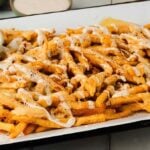 Crispy fries, known as Louisiana voodoo fries, are topped with melted cheese and drizzled with white sauce, served on a tray alongside a small dish of ketchup.