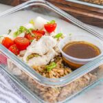 Glass container with meal prep Caprese chicken, cooked rice, cherry tomatoes, and onions. A small cup of dark dressing is included. A striped cloth is partially visible beside the container.