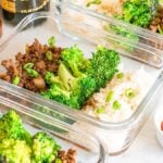 Glass containers with meal prep firecracker beef, cooked rice, broccoli, and green onions. A small dish of spicy red sauce is ready to ignite your senses. A striped cloth rests beside them.