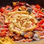 A pan of shakshuka with a swirl of hummus and sprinkled pine nuts in the center, reminiscent of a Mediterranean chickpea skillet. Surroundings include tomatoes, eggplant, bell peppers, and herbs.
