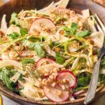 Fresh salad with shaved asparagus, radishes, greens, fennel, and nuts in a wooden bowl, served with utensils and a lemon wedge nearby.