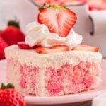 A slice of strawberry poke cake topped with whipped cream and fresh strawberries sits invitingly on a pink plate.