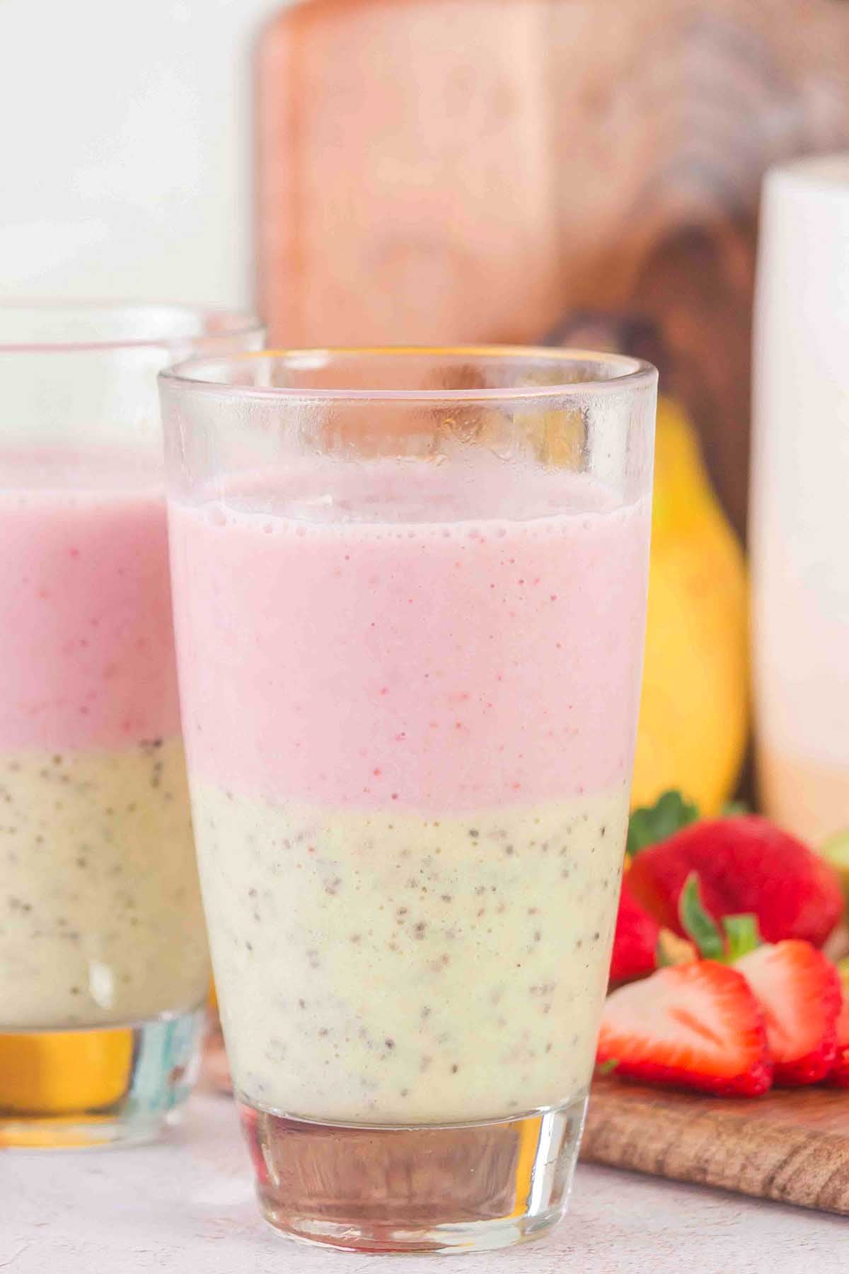 A glass of Layered Kiwi Strawberry Smoothie with chia seeds, next to fresh strawberries and a banana.