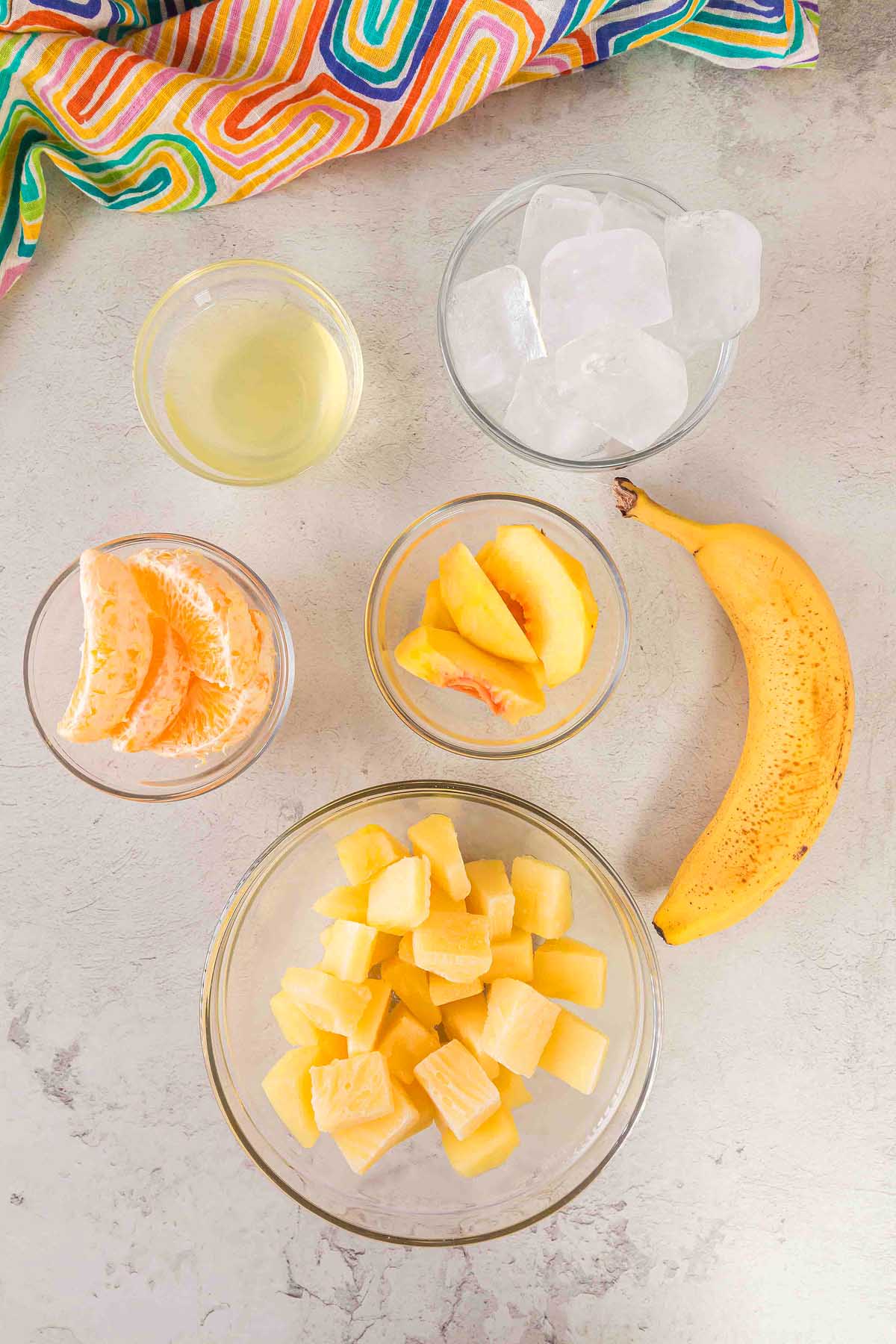 Five bowls with fruit—pineapple, orange, peach—banana, ice, and juice are arranged on a light surface with a colorful cloth, perfect for creating a refreshing Pineapple Peach Smoothie.