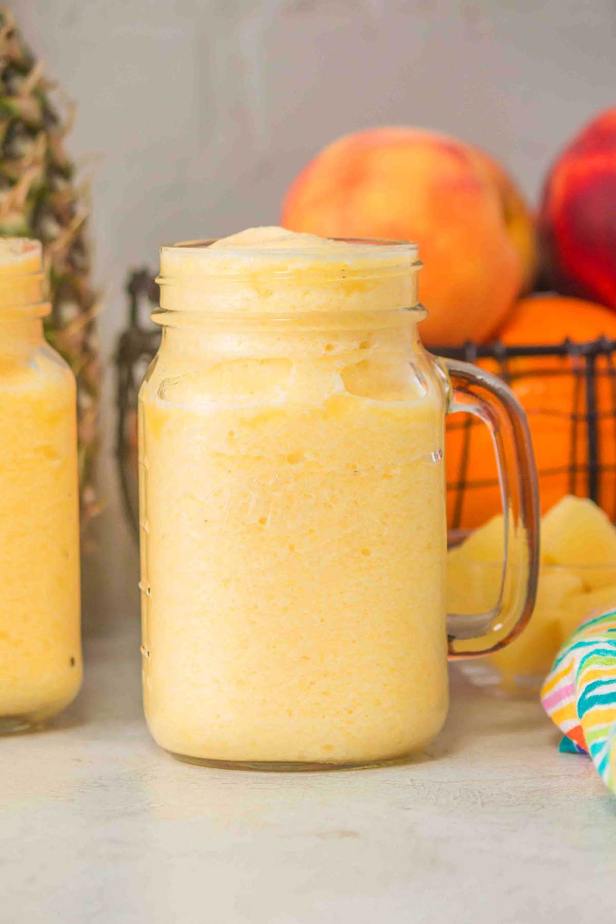A frothy yellow Pineapple Peach Smoothie in a mason jar, with juicy peaches, fresh pineapple, and vibrant orange in the background.