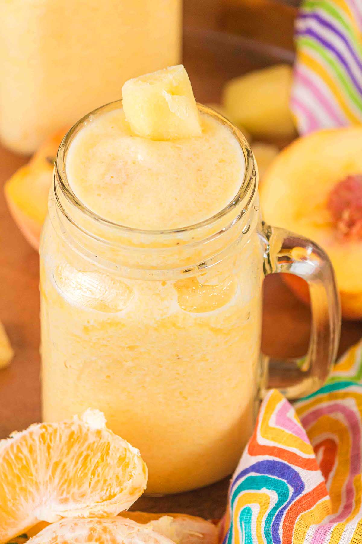 A mason jar filled with Pineapple Peach Smoothie, topped with pineapple chunks, sits next to orange slices and a colorful cloth.