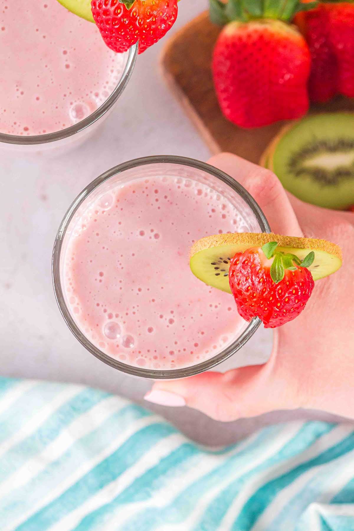 A hand holds a Layered Kiwi Strawberry Smoothie, garnished with a kiwi slice and strawberry, beside fresh fruit.