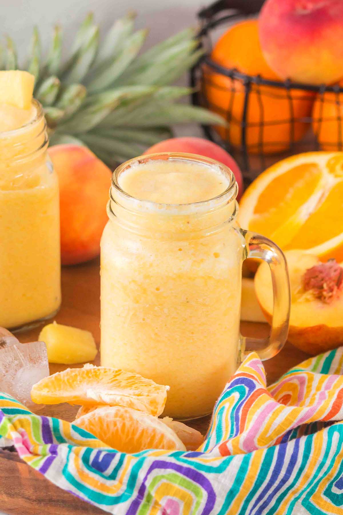 A mason jar filled with a creamy Pineapple Peach Smoothie, surrounded by fresh oranges, peaches, and pineapple.