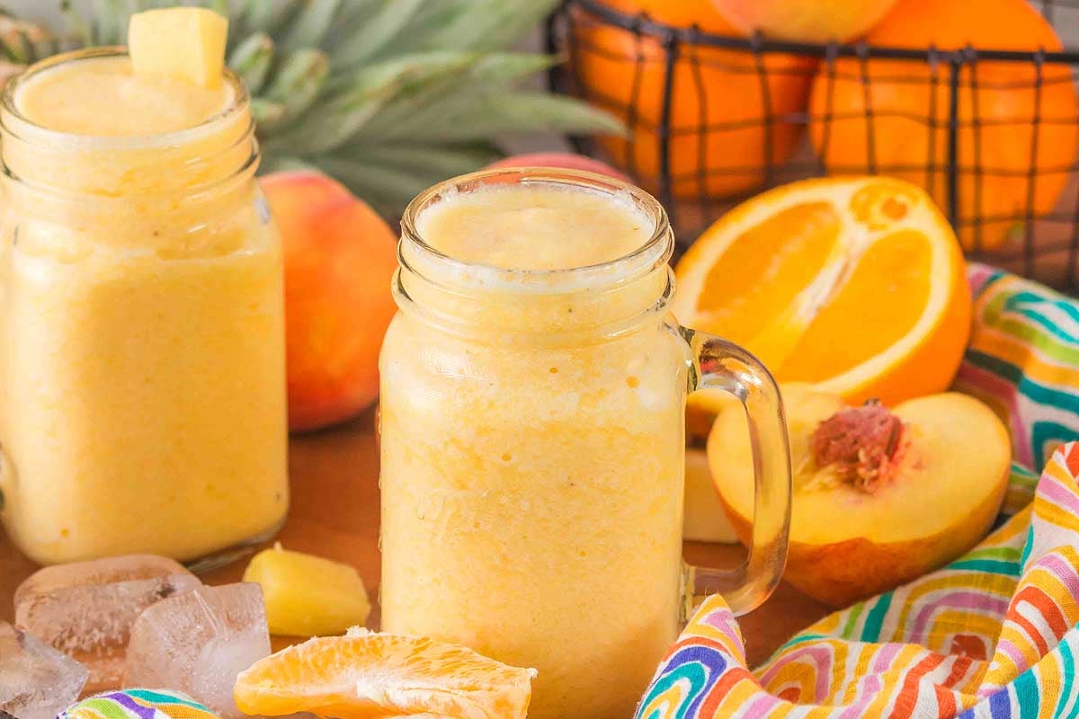 Two mason jars of Pineapple Peach Smoothie with fruit slices, ice cubes, and a colorful cloth nearby.