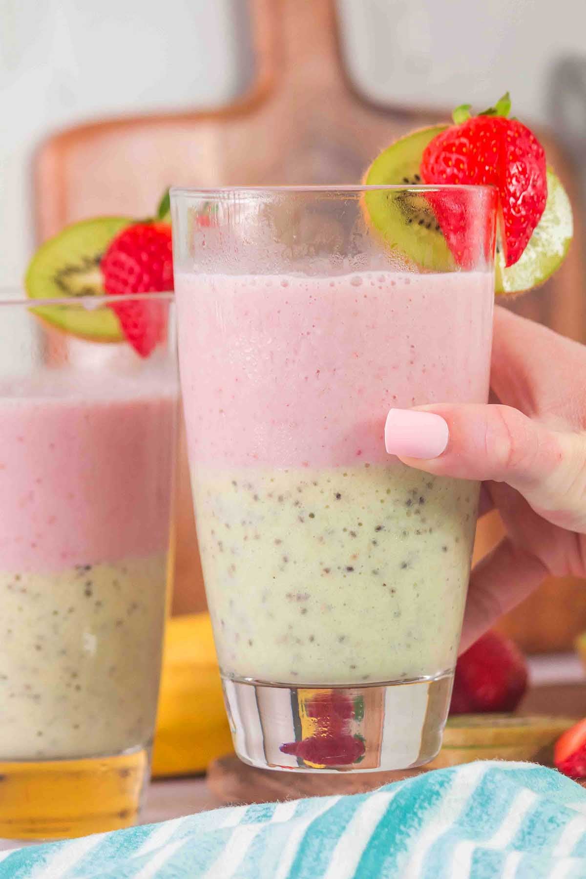 Hand holding a vibrant Layered Kiwi Strawberry Smoothie with pink and green sections, garnished with fresh kiwi and strawberry slices.