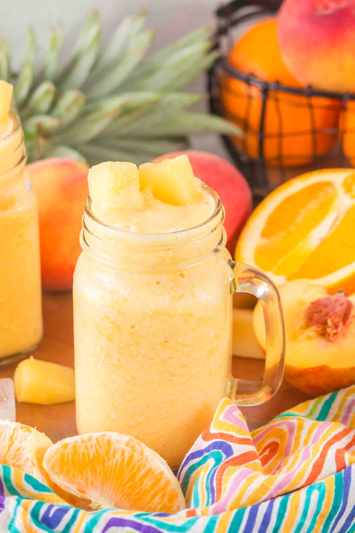 A mason jar filled with a yellow Pineapple Peach Smoothie, topped with pineapple, surrounded by fresh fruit and a colorful cloth.