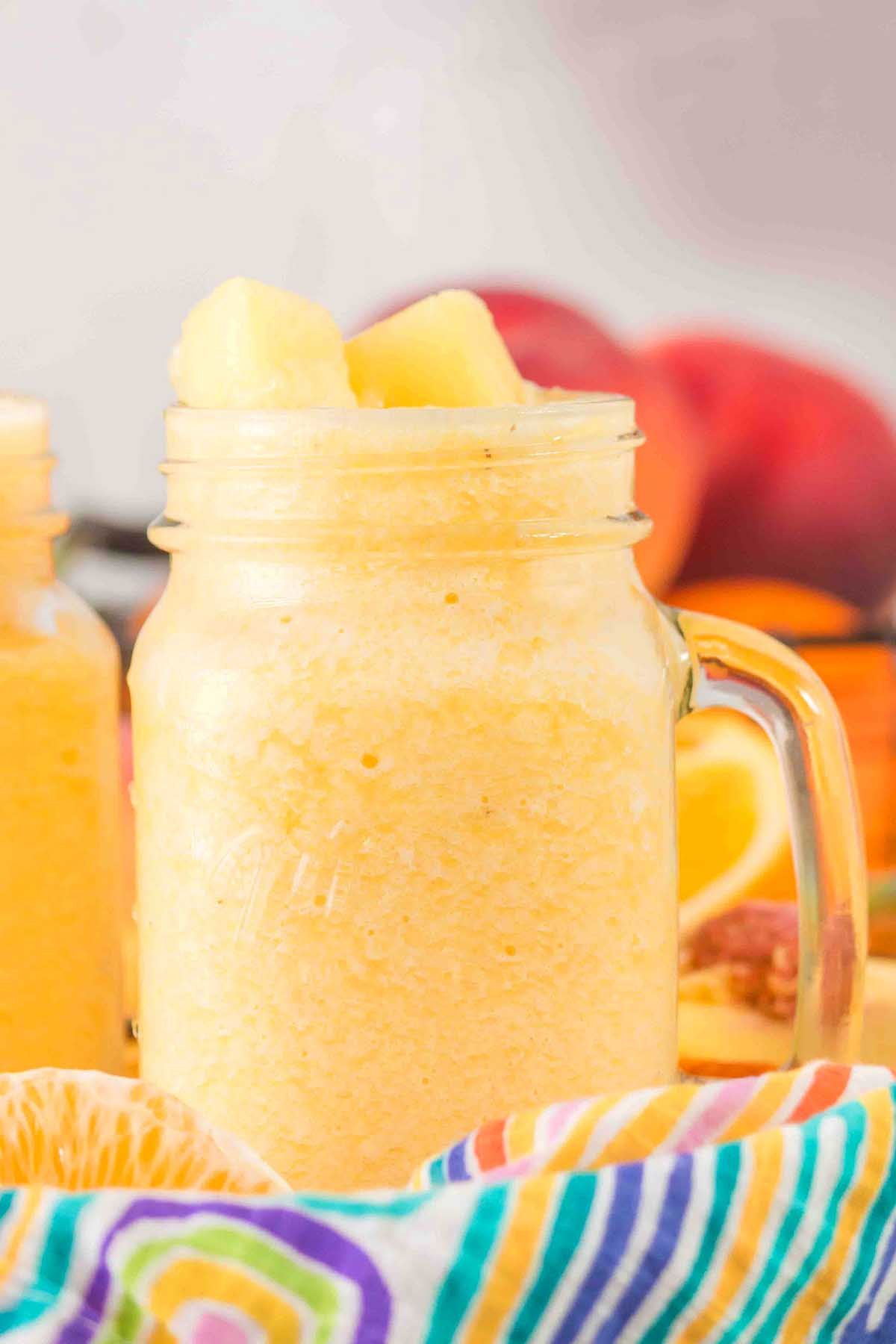 A mason jar filled with a Pineapple Peach Smoothie, topped with pineapple chunks, with fresh fruit in the background.