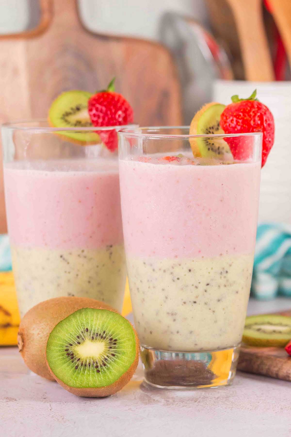 Two glasses of Layered Kiwi Strawberry Smoothie, topped with kiwi and strawberry slices, sit alongside fresh fruit for a vibrant and refreshing treat.