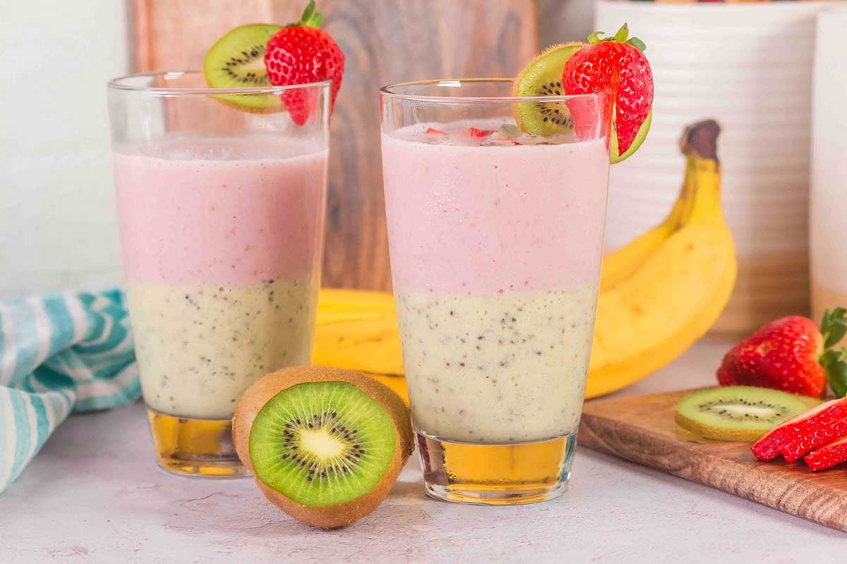 Two glasses of Layered Kiwi Strawberry Smoothie are garnished with strawberries, with fresh fruit arranged beside them.