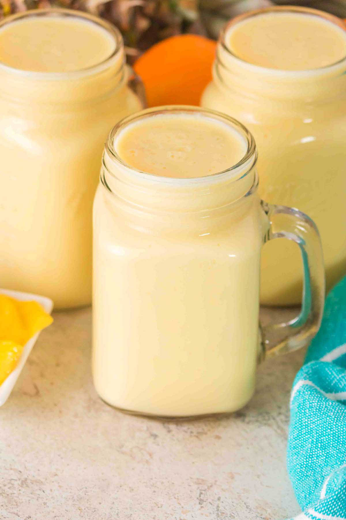 A mason jar filled with creamy, pale yellow Pineapple Mango Smoothie sits on a counter next to two similar jars.