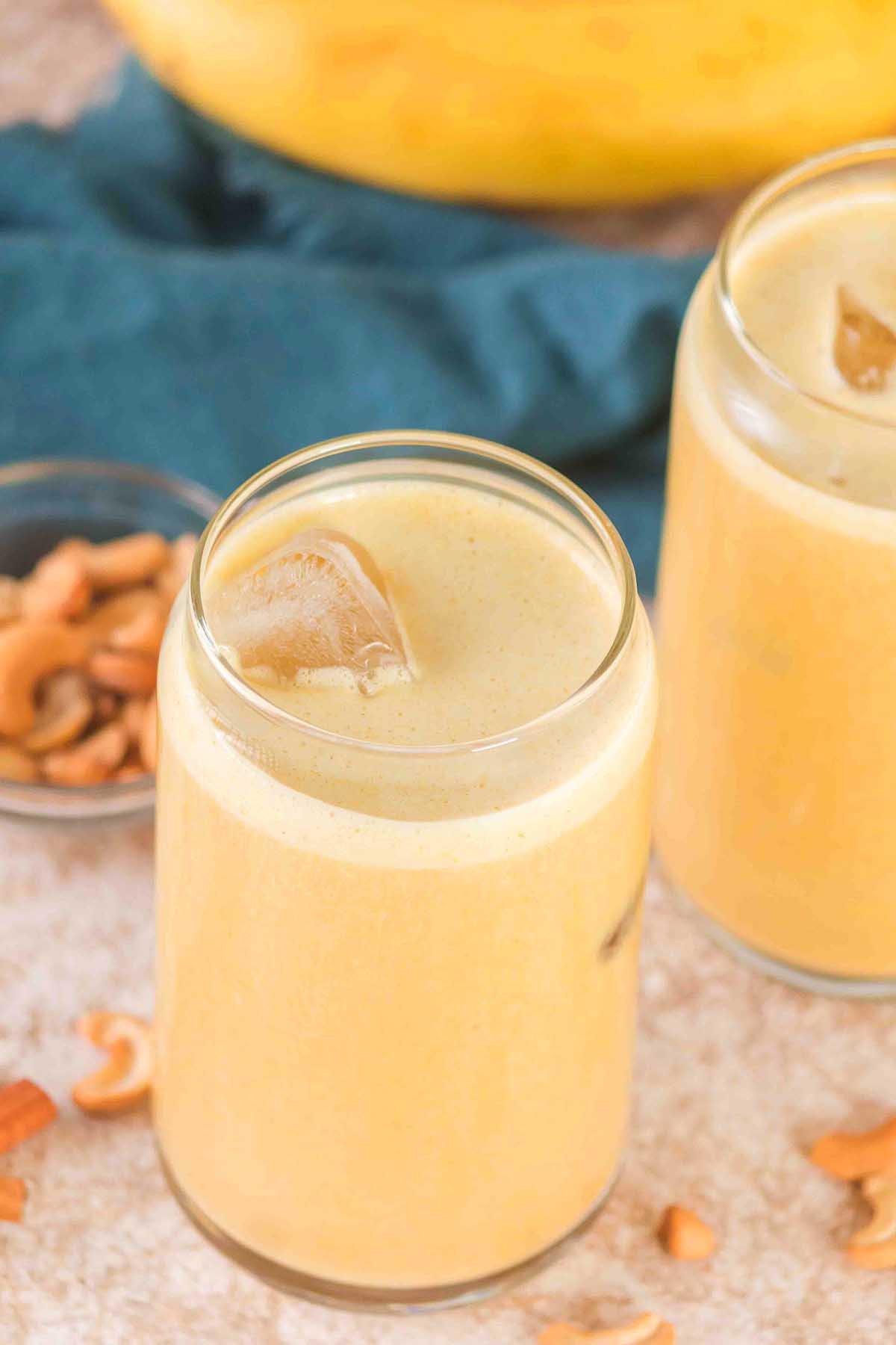 Two glasses of creamy yellow Banana Cashew Smoothie with ice cubes, surrounded by scattered cashews on a light surface.