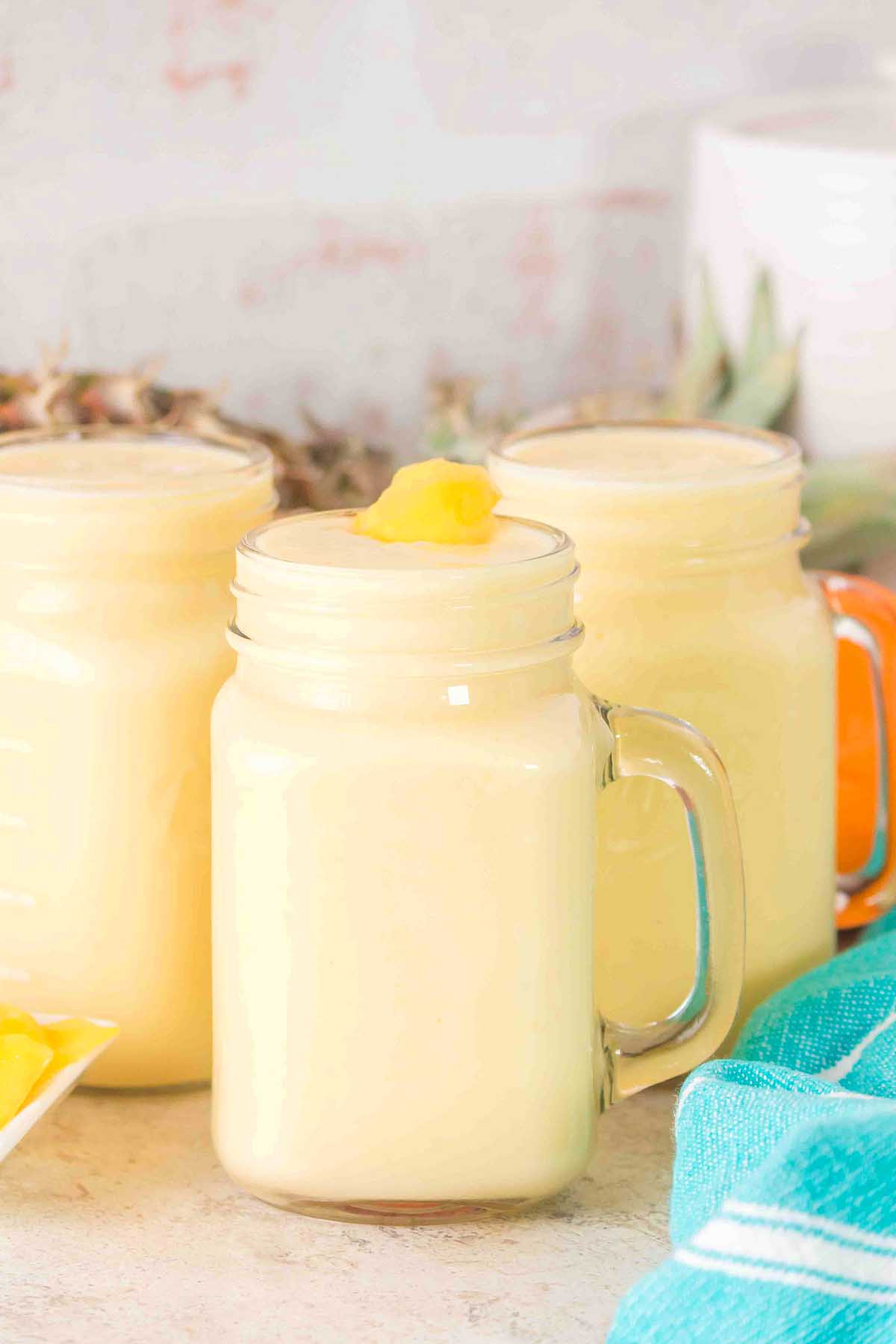 Three glass mugs filled with creamy yellow Pineapple Mango Smoothie, each topped with a fresh pineapple chunk.