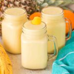 Three mason jars filled with Pineapple Mango Smoothie, surrounded by bananas, pineapples, and an aqua towel.