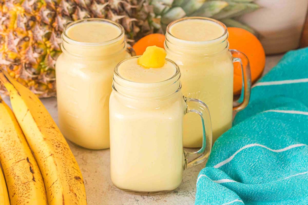 Three mason jars filled with Pineapple Mango Smoothie, surrounded by bananas, pineapples, and an aqua towel.