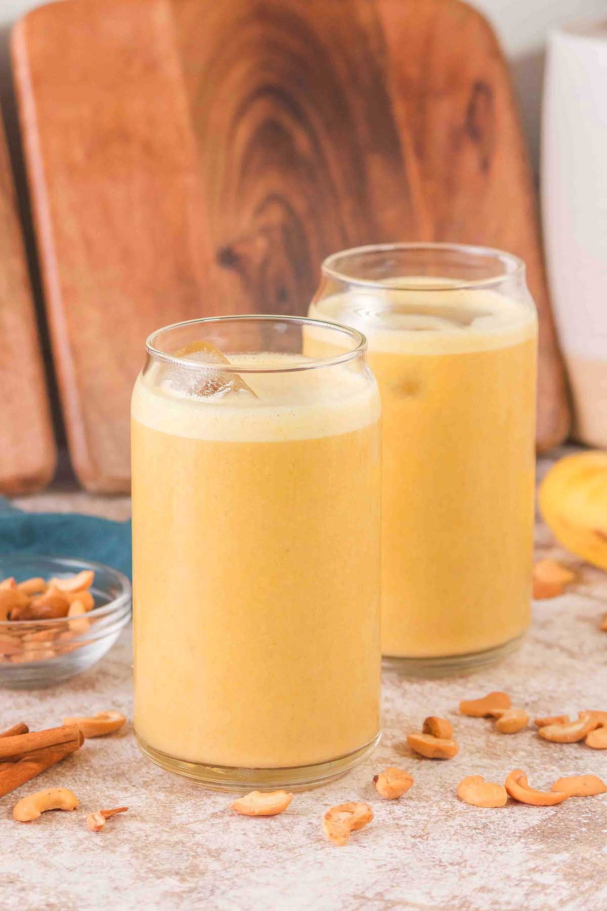 Two glasses of creamy yellow Banana Cashew Smoothie with ice, surrounded by cashews and a cinnamon stick on the counter.