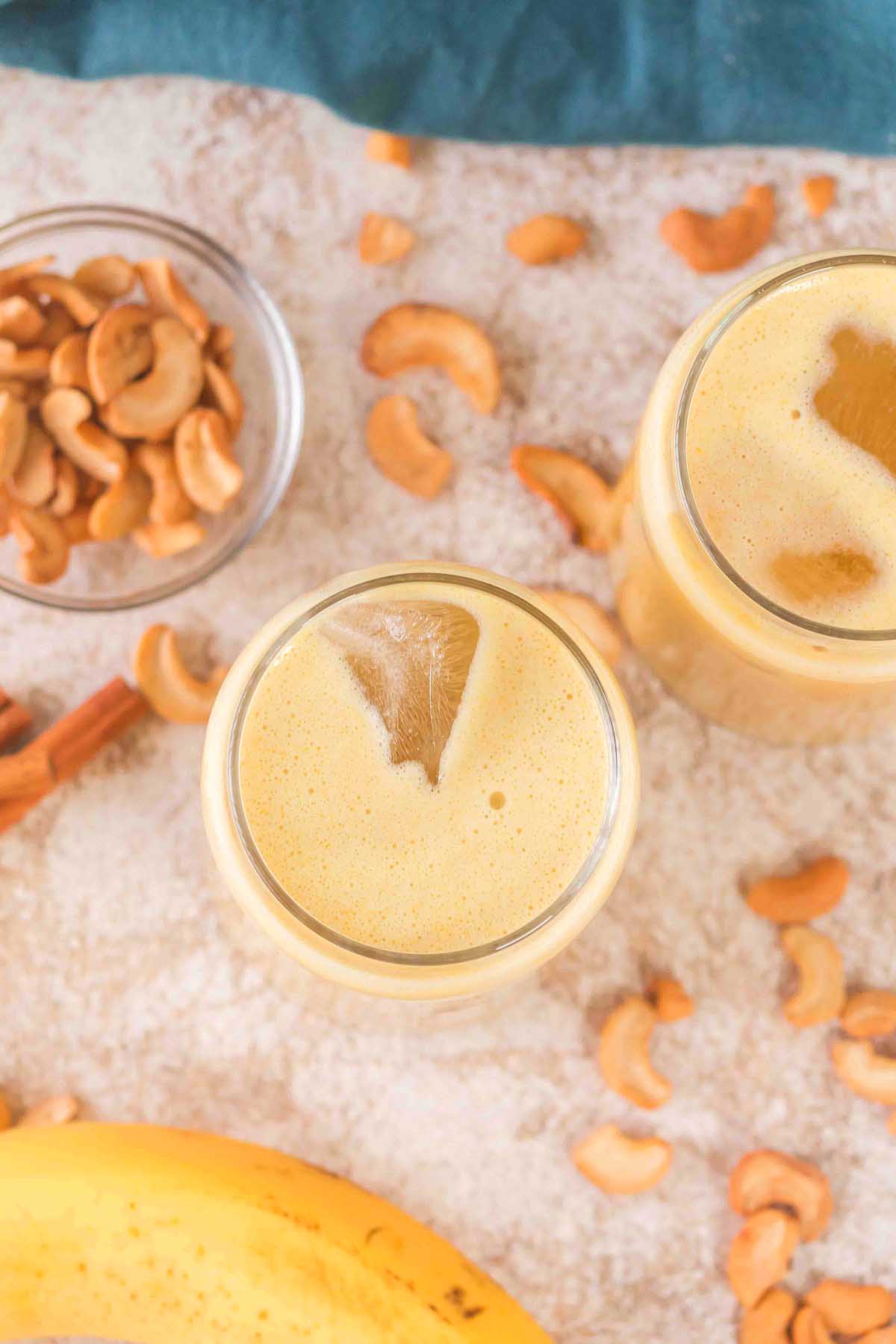 Overhead view of two glasses of creamy Banana Cashew Smoothie, with cashews and banana slices scattered around.