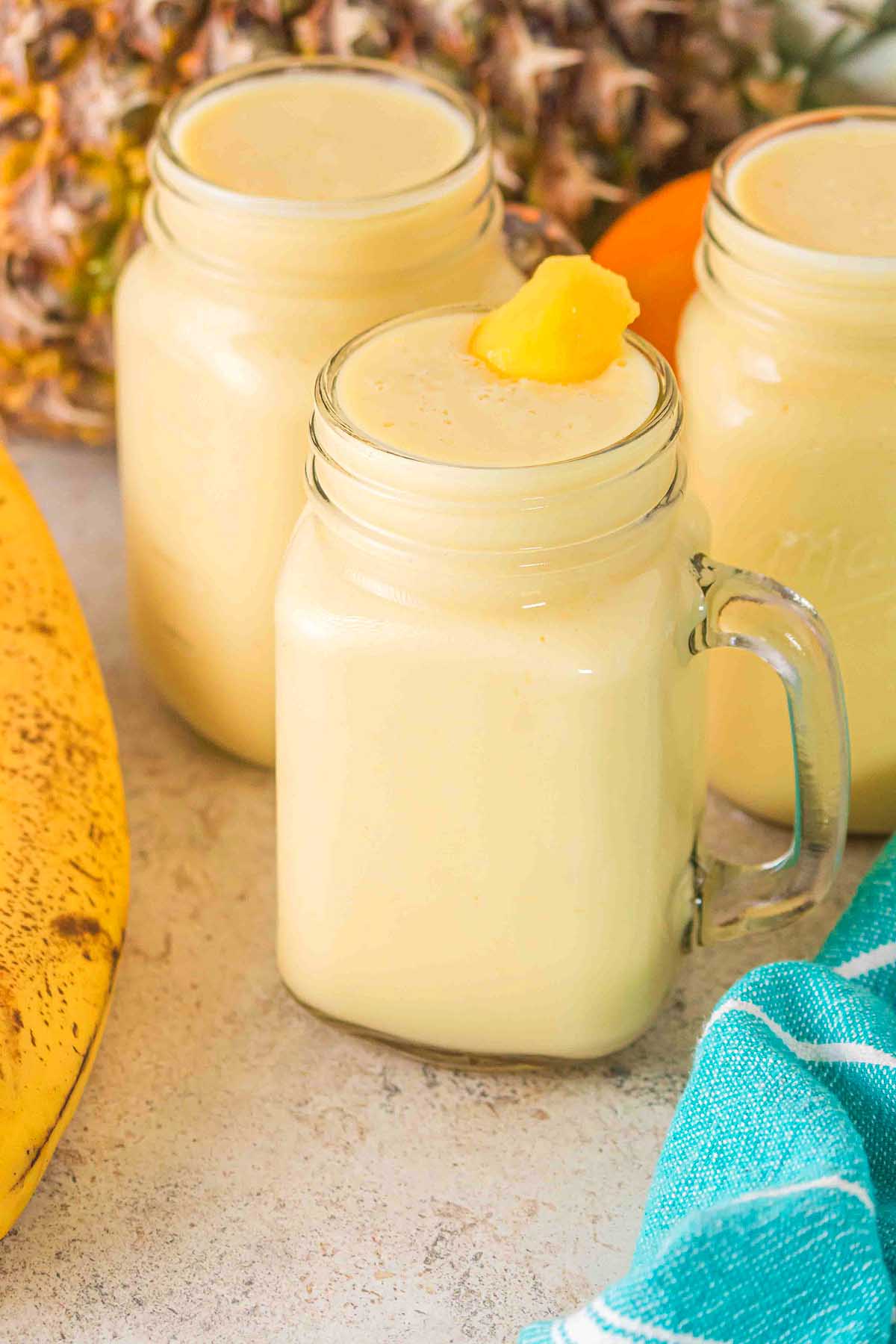 Three mason jars filled with creamy yellow Pineapple Mango Smoothie, one topped with pineapple, sit next to a banana and a blue cloth.
