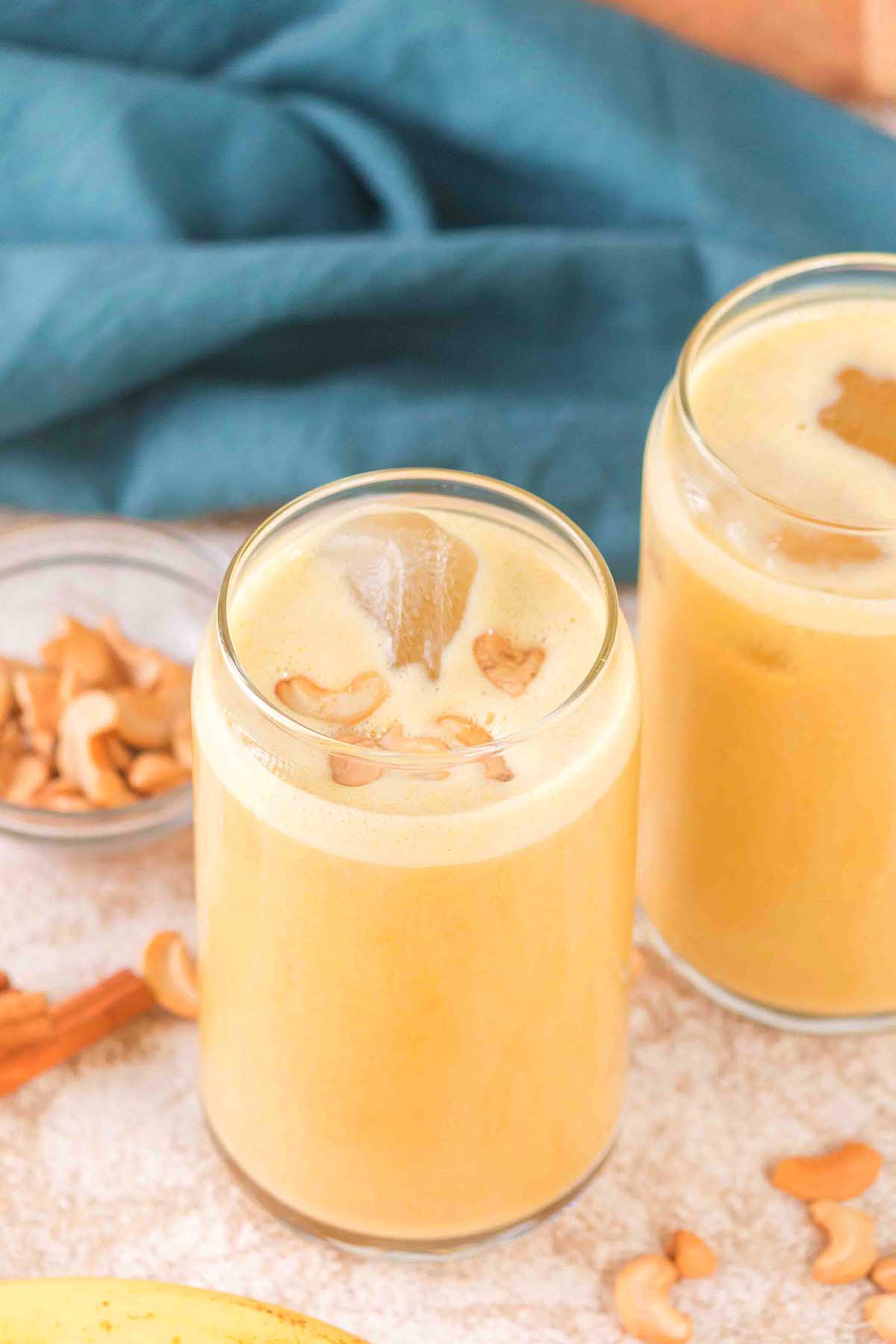 Two glasses of creamy yellow Banana Cashew Smoothie topped with cashew pieces, with cashews and cinnamon nearby.