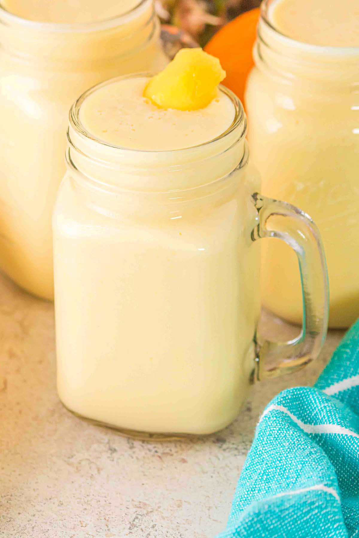 A mason jar filled with creamy yellow Pineapple Mango Smoothie, topped with a pineapple chunk, on a light surface.